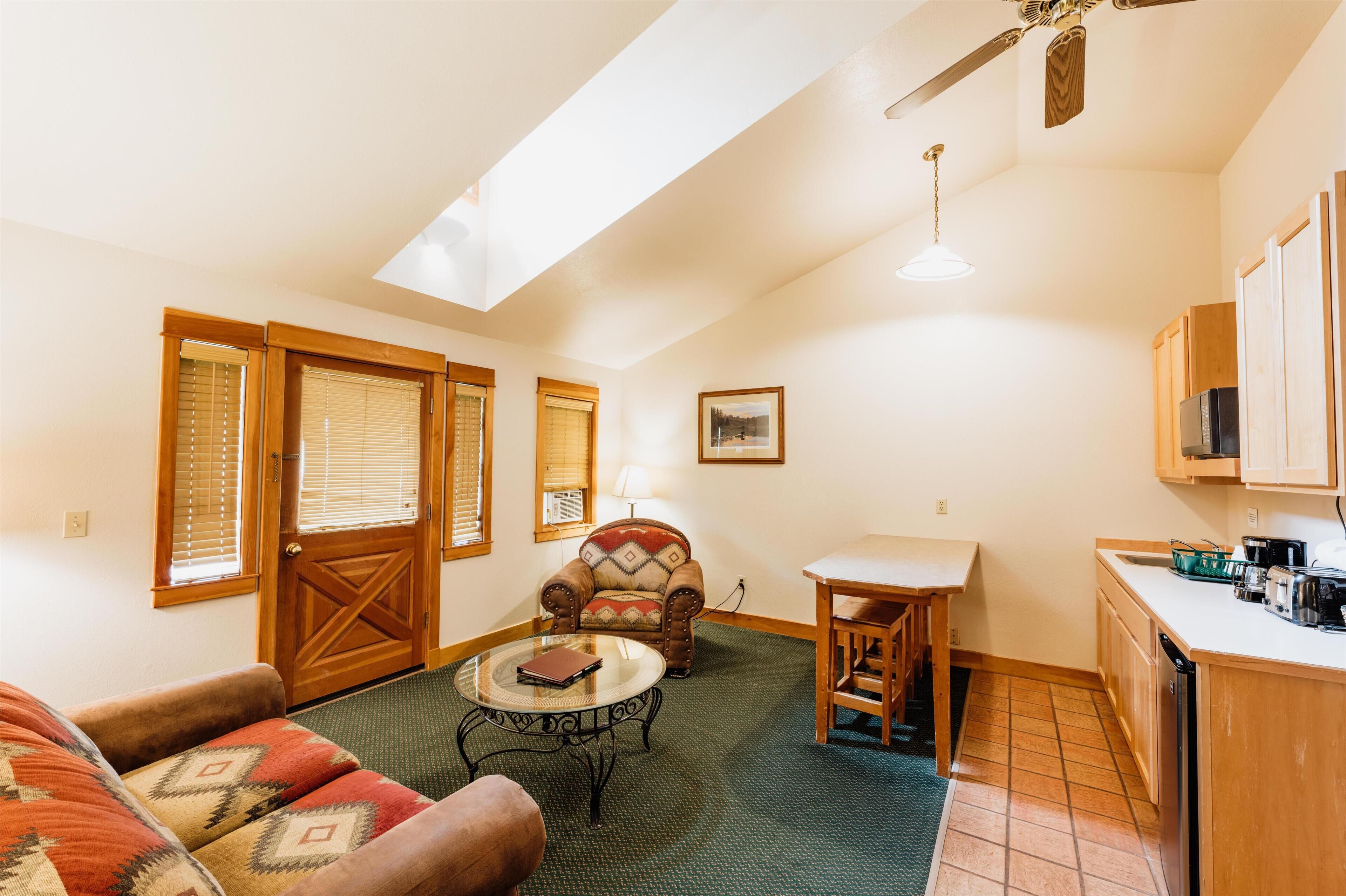 A cozy living room with a sofa, armchair, coffee table, and a dining table. There is a kitchen area with a refrigerator and cabinets. A ceiling fan and a window with blinds are also visible.