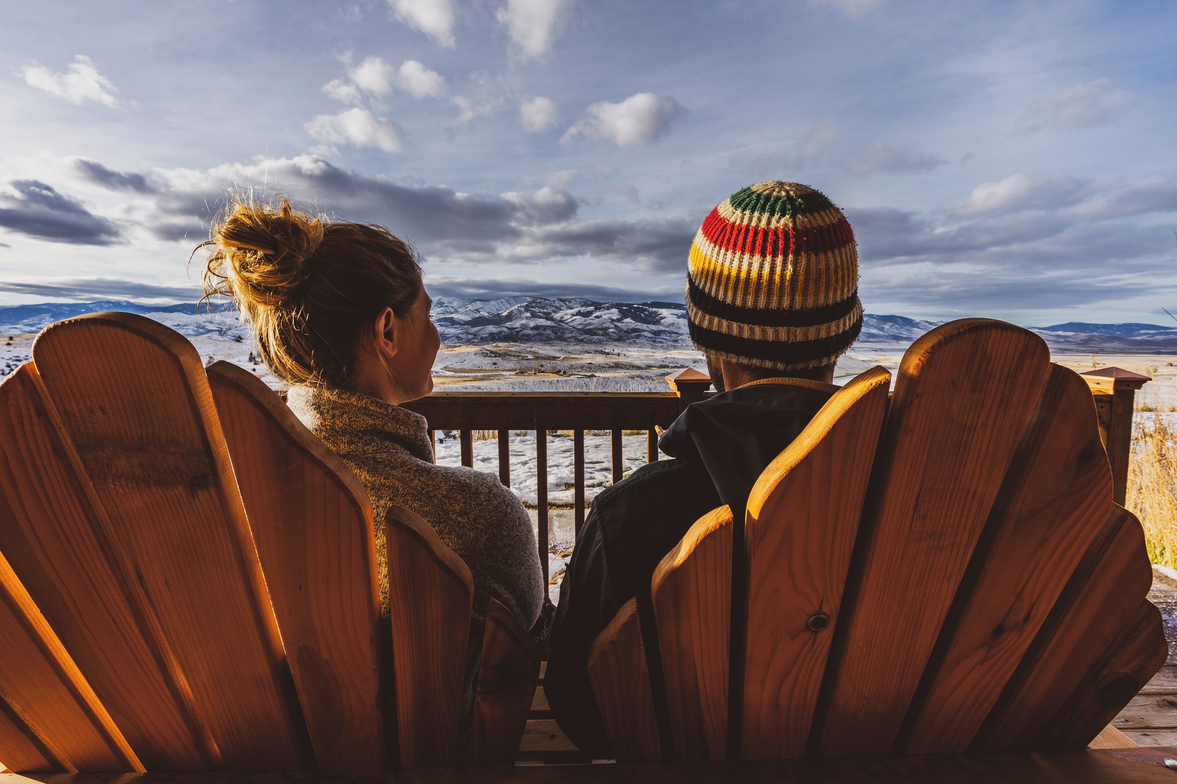 Two people sitting on wooden chairs facing a snowy landscape with mountains in the background.