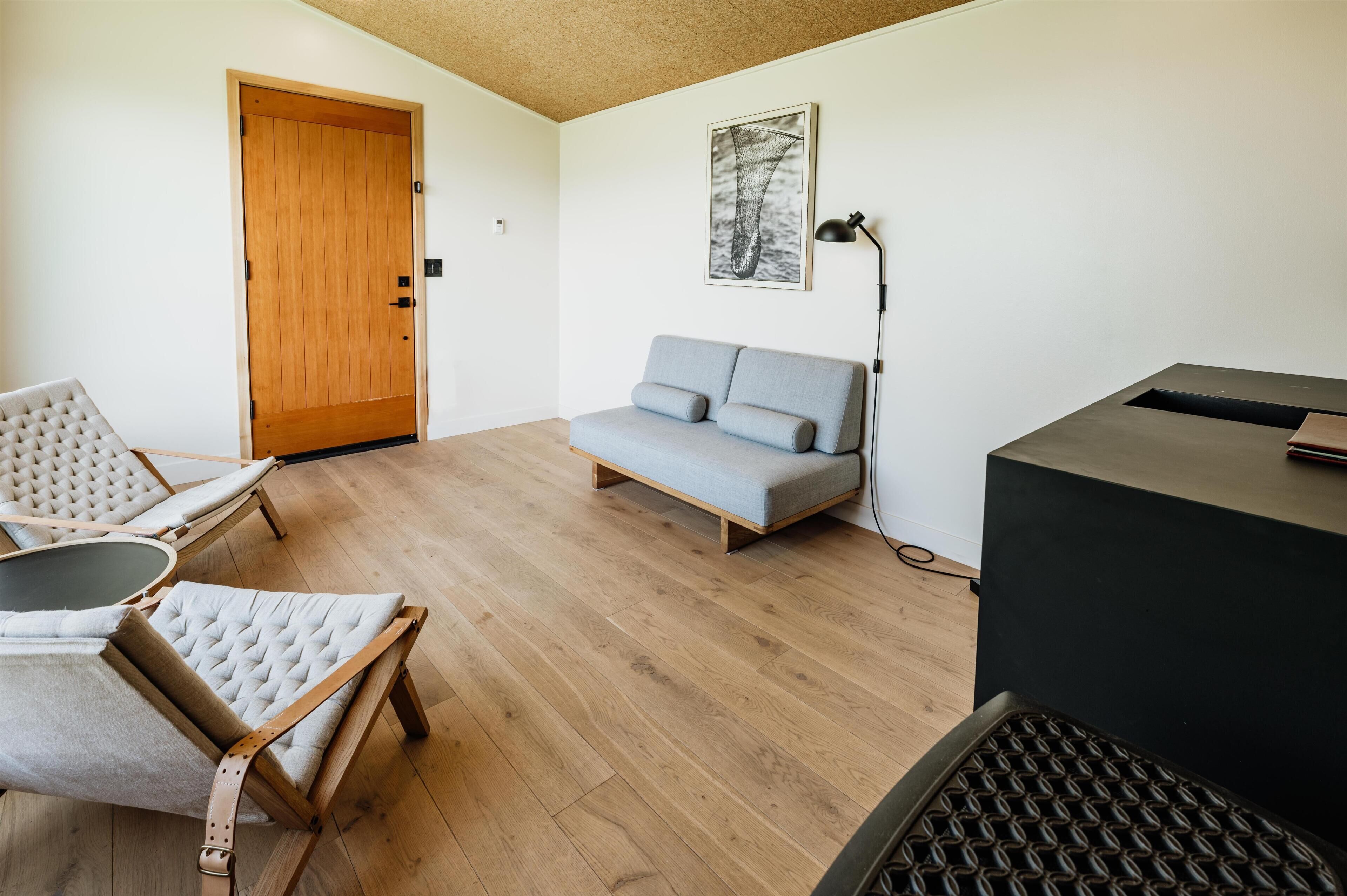 Ein Zimmer mit Holztür, grauem Sofa, zwei Sesseln, einem schwarzen Schreibtisch und einem gerahmten Bild an der Wand.
