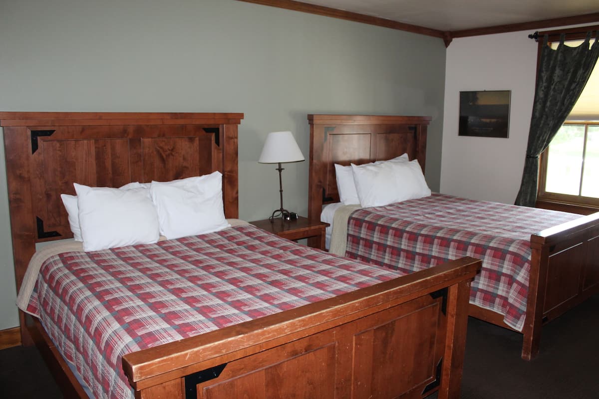A room with two wooden beds, each with a plaid bedspread and white pillows. There is a nightstand with a lamp between the beds.
