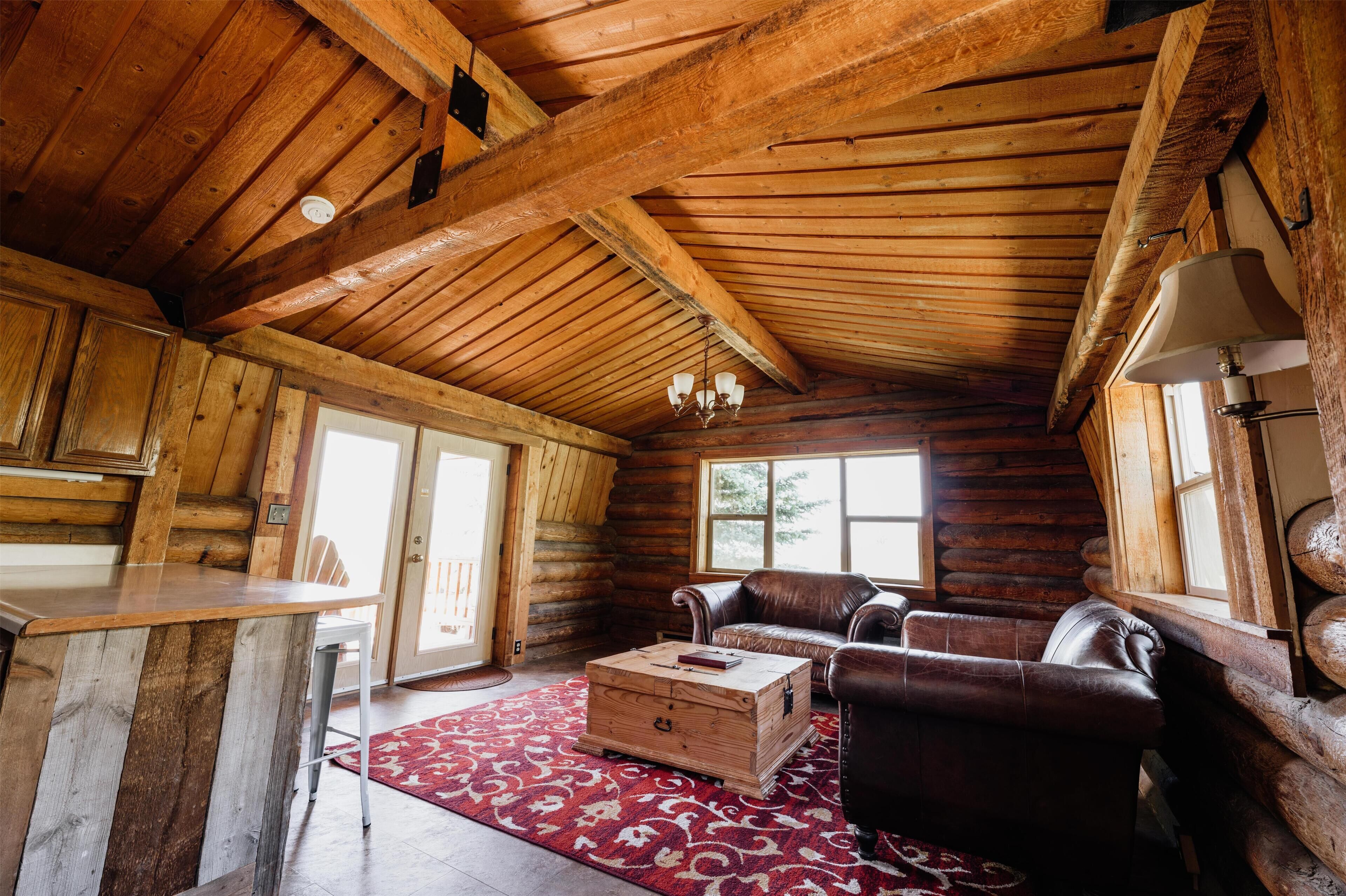 A log cabin interior with a wooden ceiling, a dining area with a bar counter, a leather sofa, and a red patterned rug.