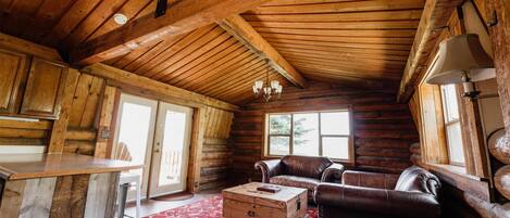 A log cabin interior with a wooden ceiling, a dining area with a bar counter, a leather sofa, and a red patterned rug.