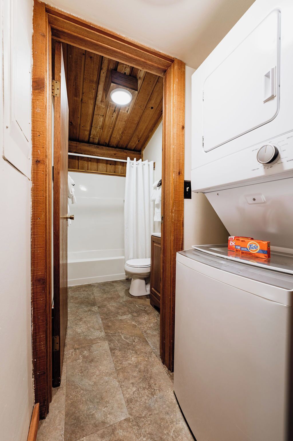 A compact bathroom with a wooden ceiling, a white toilet, a shower with a white curtain, and a washing machine with Tide detergent.