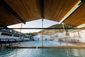 A swimming pool area with a wooden roof, a view of a building with 'HICO' signage, and a mountainous backdrop. - Chico Hot Springs Resort & Day Spa (Pray)