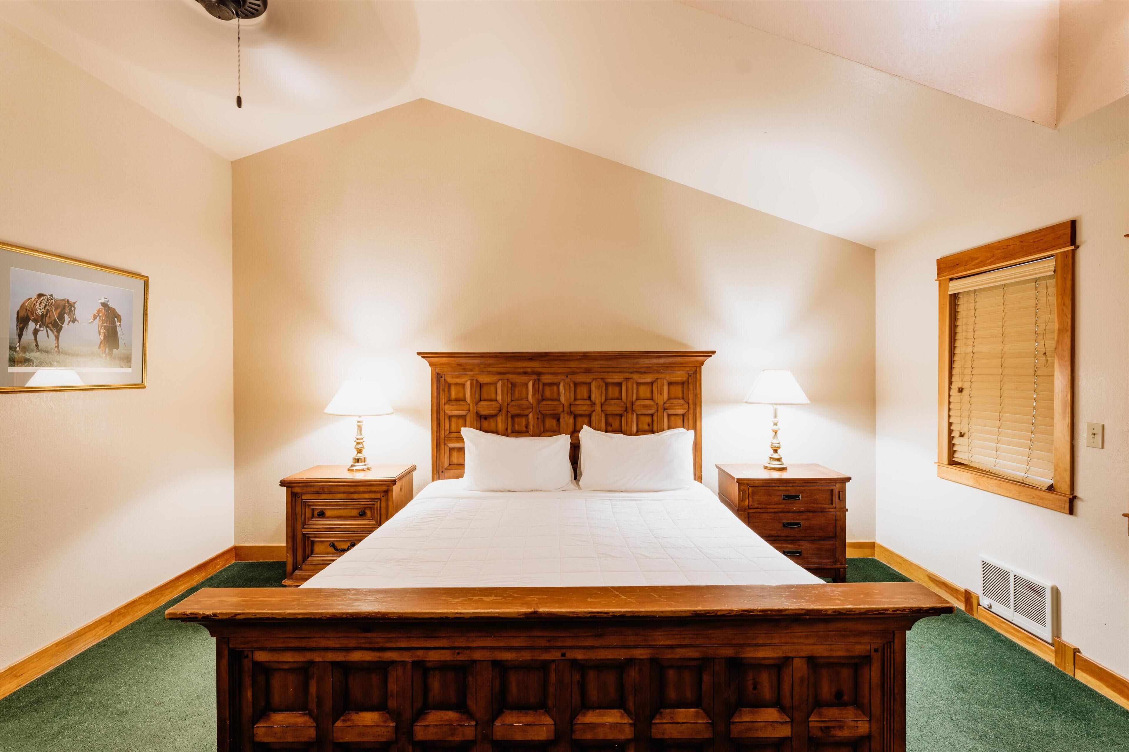 Un dormitorio con una cama de madera, dos mesitas de noche con lámparas, un cuadro en la pared y una ventana con persianas.