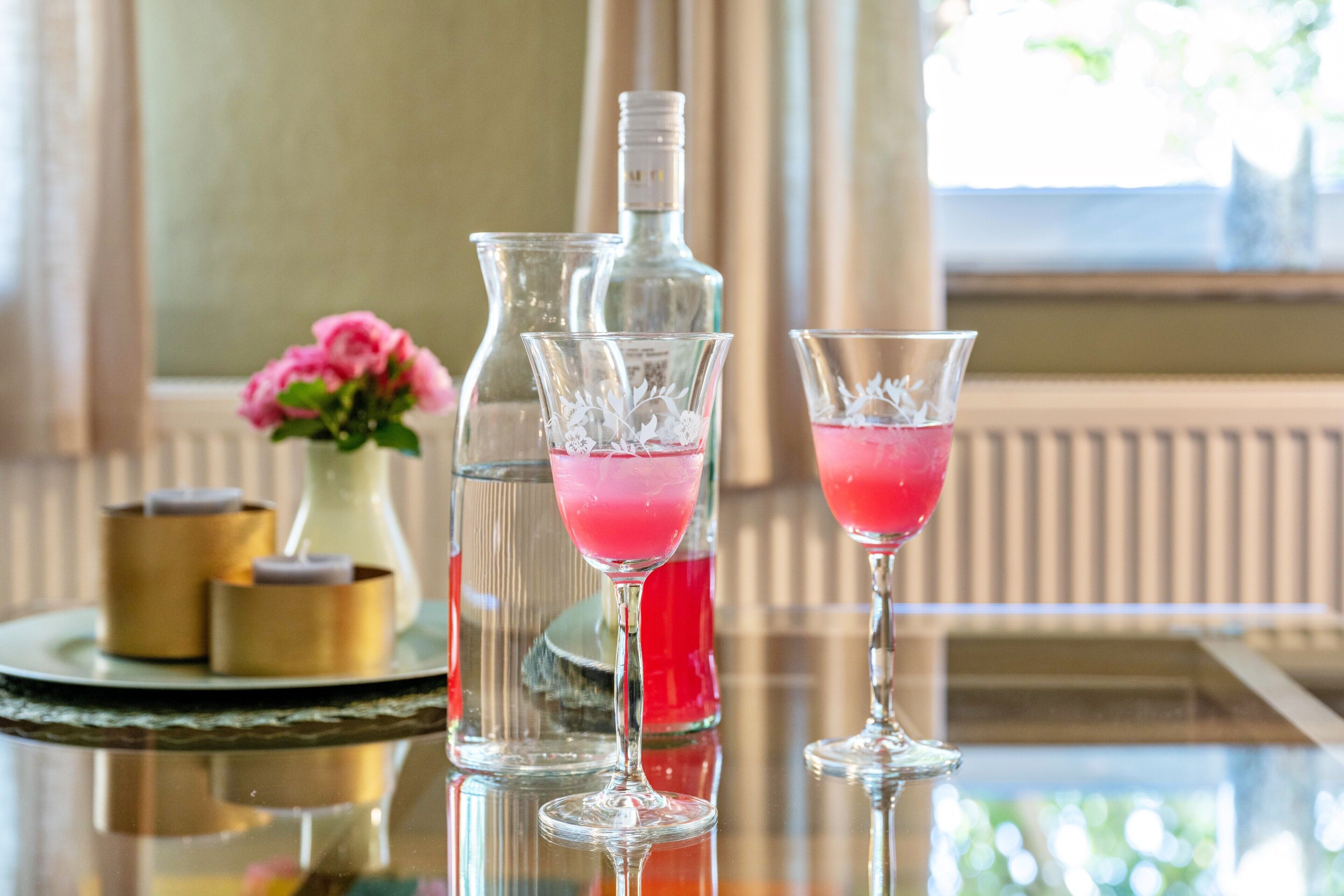 Un tavolo con una bottiglia di vetro, due bicchieri con bevande rosa e un vaso con fiori rosa.