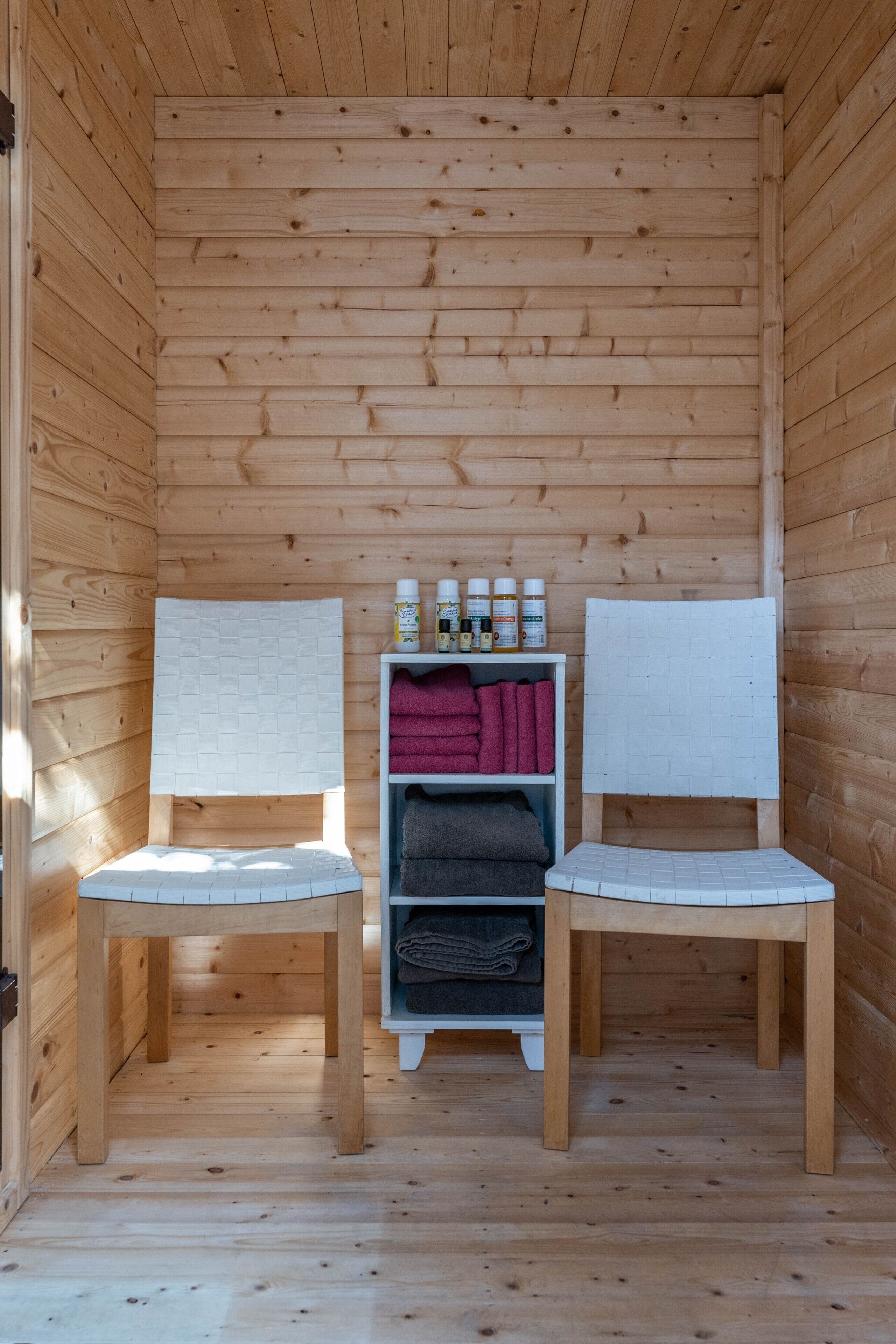 Une petite cabane en bois avec deux chaises blanches, une étagère contenant des serviettes et des articles de toilette, et un plancher en bois.