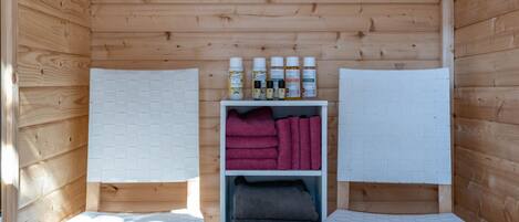 Une petite cabane en bois avec deux chaises blanches, une étagère contenant des serviettes et des articles de toilette, et un plancher en bois.