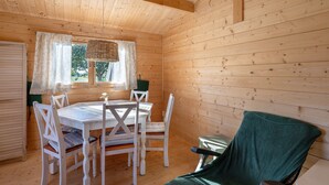 Interior de una cabaña de madera con una mesa de comedor, sillas blancas, una silla verde, una ventana con cortinas y una lámpara colgante.