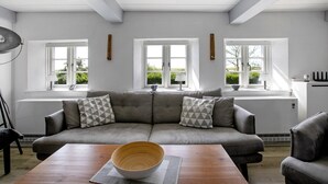 A modern living room with a grey sofa, two windows, and a wooden table. - Holiday Home 'Pellworm-Kate' with Private Terrace, Private Garden and Wi-Fi (Pellworm)