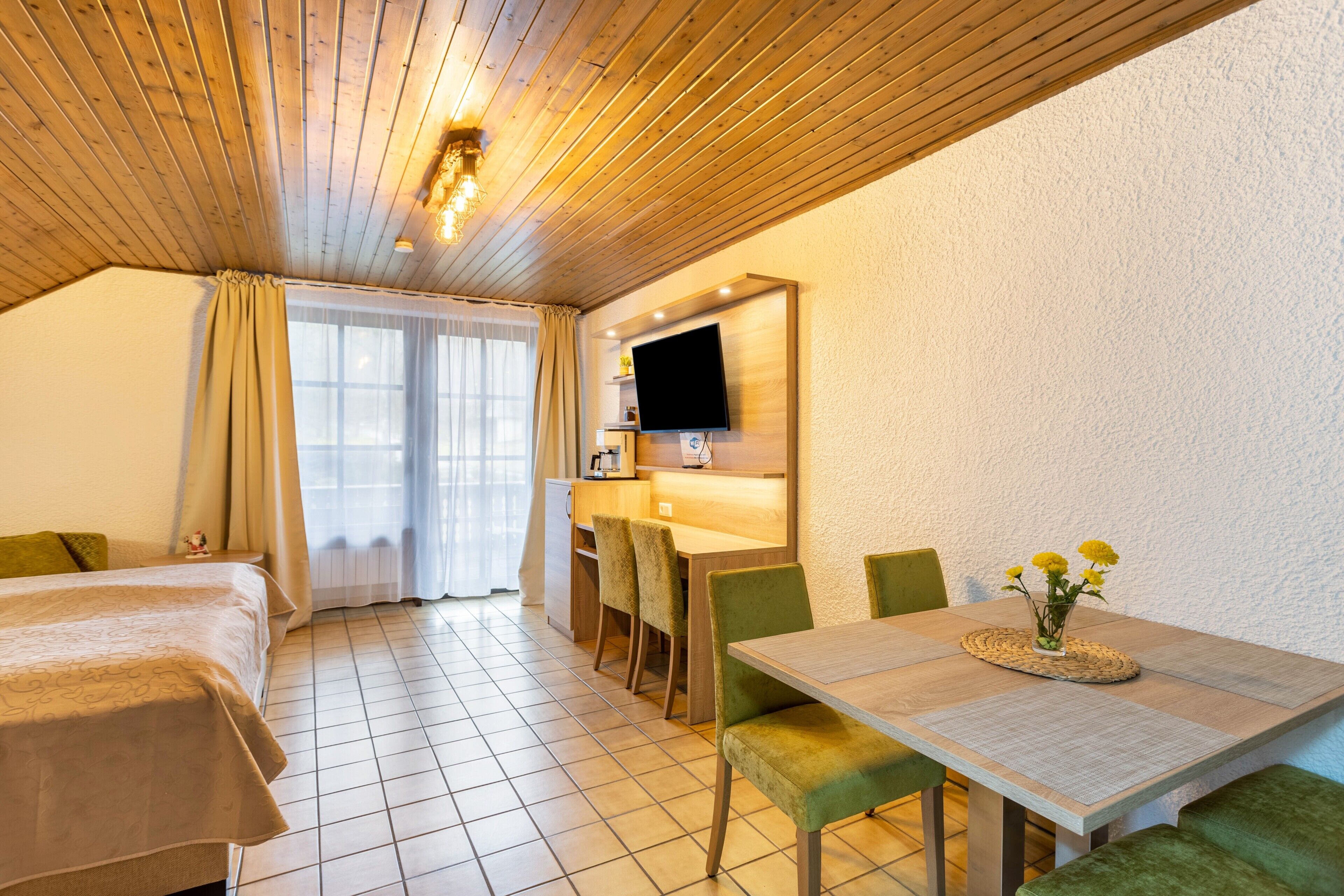 Ein Zimmer mit einem Bett, einem Esstisch mit Stühlen, einem an der Wand befestigten Fernseher und einem Fenster mit Vorhängen.