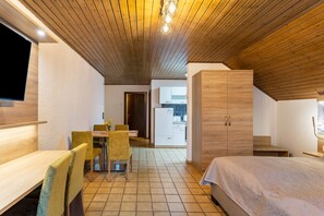 A compact room with a bed, a desk, a chair, a dining table with chairs, a kitchenette, and a TV mounted on the wall.