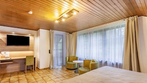 Habitación de hotel con techo de madera, una cama, un escritorio con televisor de pantalla plana, dos sillas y un ventanal con cortinas.