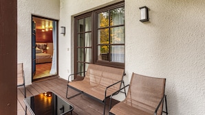 Un patio techado con un piso de madera, dos sillas de mimbre, una mesita y una habitación visible a través de una puerta abierta.