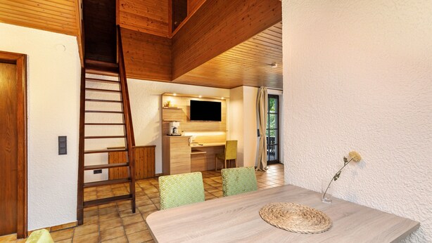 Un comedor con una mesa de madera, sillas, un mantel tejido, un televisor y una zona de cocina con un fregadero y armarios.
