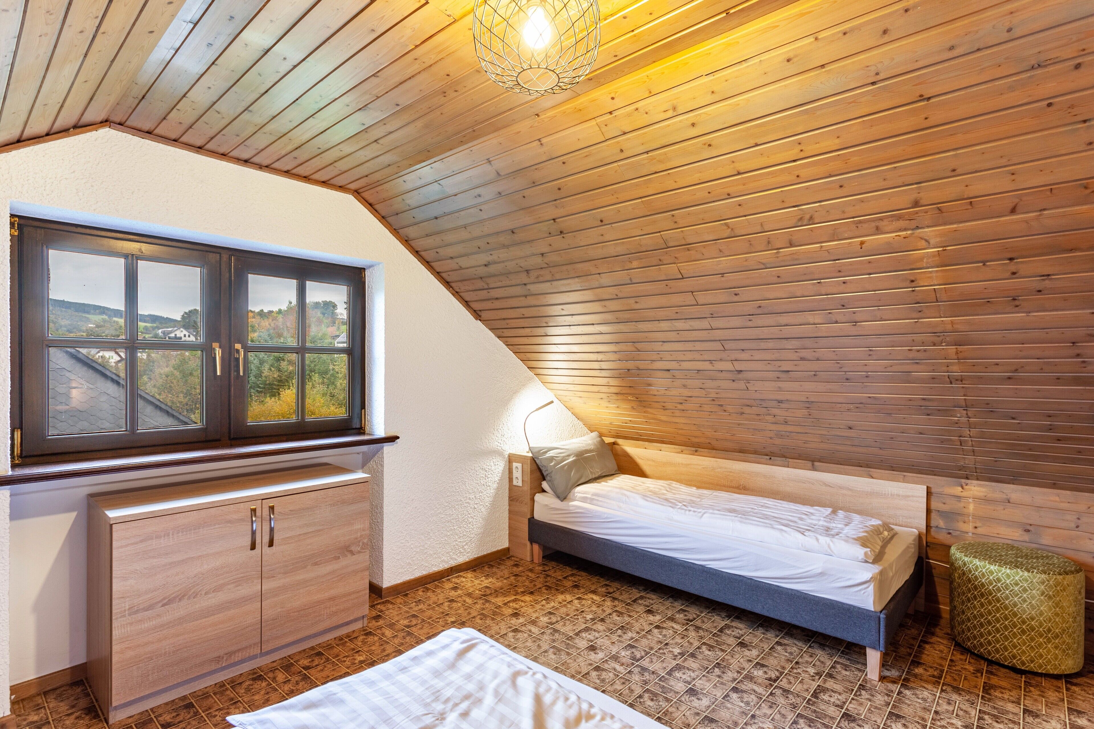 A bedroom with a sloped ceiling, a bed, a window, a cabinet, and a small stool.