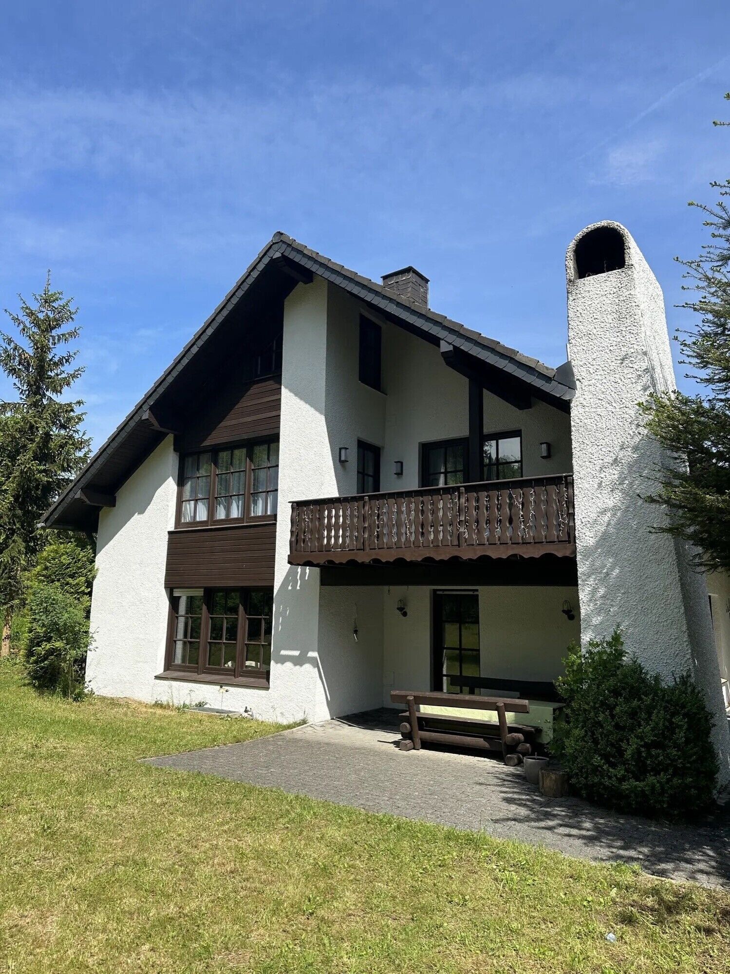 A two-story house with a balcony, a chimney, and a wooden bench in the front yard.