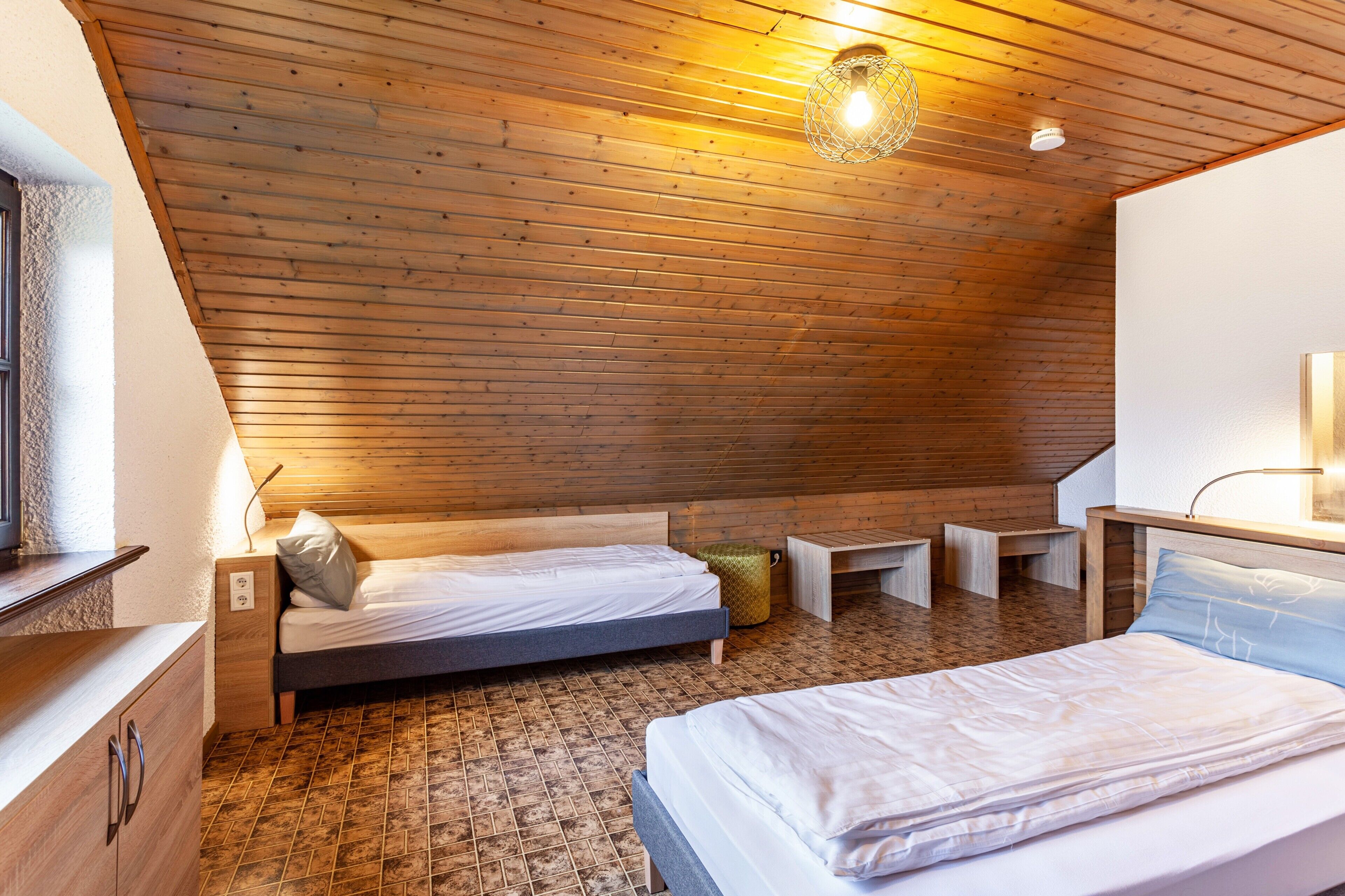 A room with wooden slatted ceiling, two beds, a small desk, and a lamp.