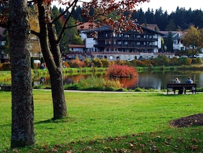 A park with a lake, trees, and a building with multiple windows and balconies.