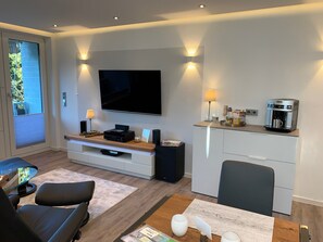 A modern living room with a flat-screen TV, a coffee machine, and a seating area with a table and chairs.