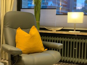 Un sillón moderno con un cojín, situado cerca de una ventana con un radiador y una lámpara sobre una mesa.