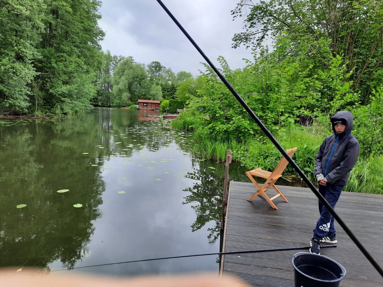 En person står på en brygga vid en stilla flod, med träd och en liten byggnad i bakgrunden.
