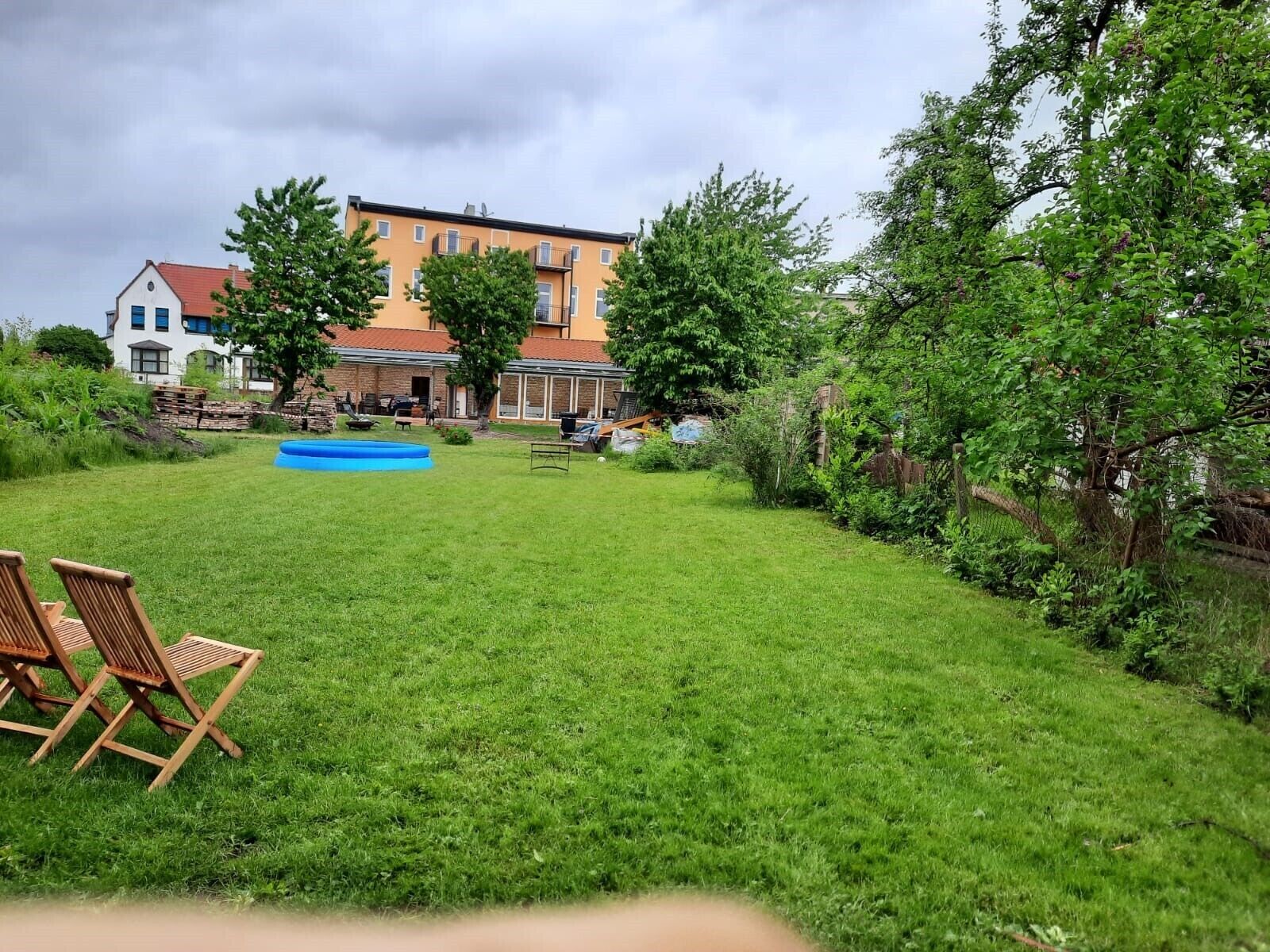 A grassy backyard with a swimming pool, wooden chairs, and a building in the background.