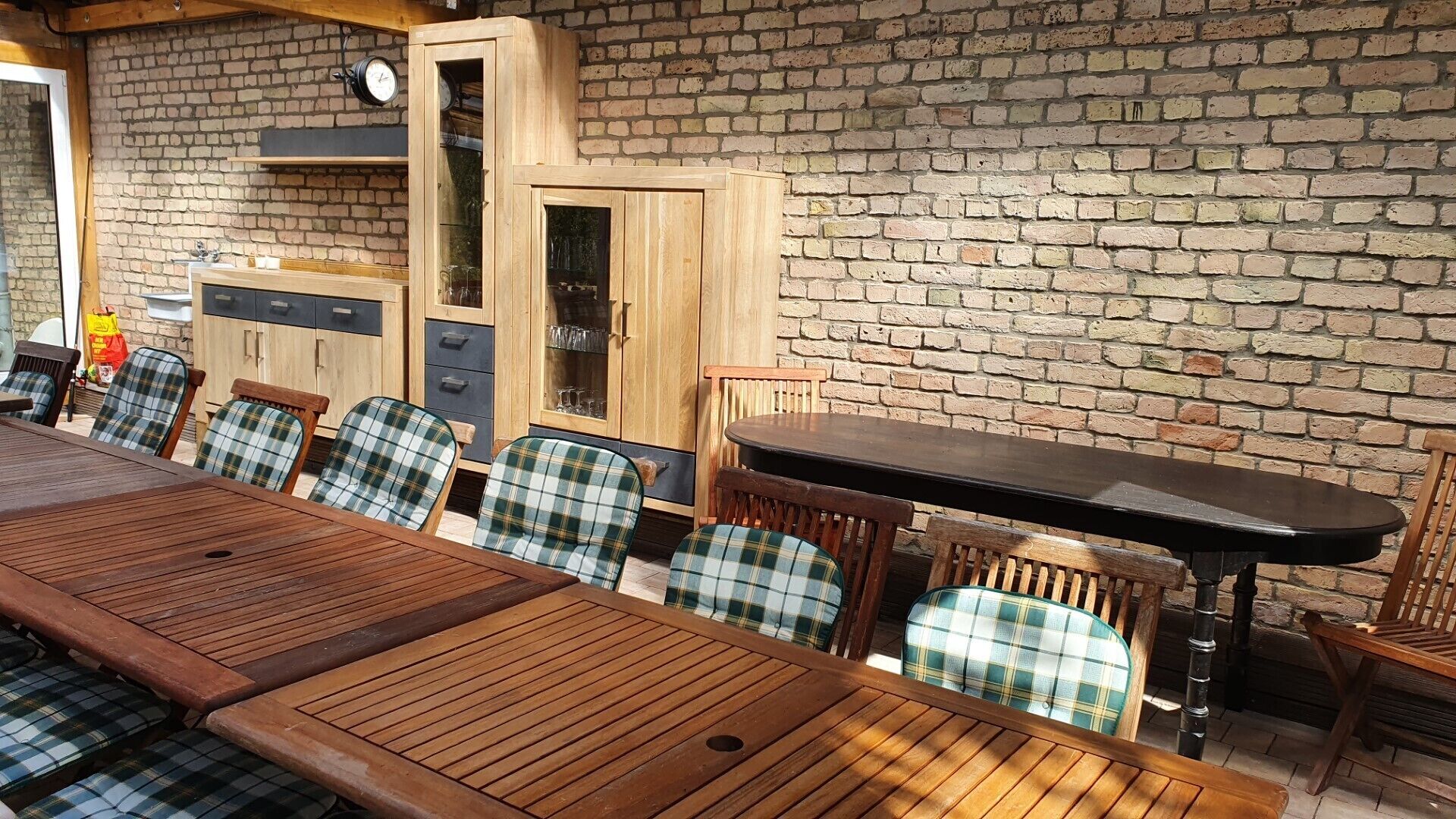 A wooden dining table with plaid chairs, a brick wall, and a wooden cabinet with glass doors.