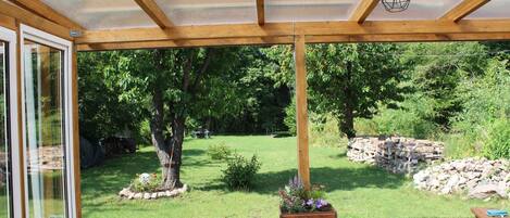 Une terrasse couverte avec un plafond en bois, un luminaire suspendu et une vue sur un jardin aménagé, agrémenté d’arbres et d’un mur en pierre.