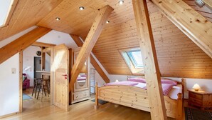 A high-ceilinged room with a sloped wooden ceiling, a bed with a checkered bedspread, a bedside table with a lamp, and a skylight.