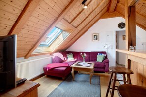Ein gemütliches Zimmer mit schrägem Dach, einer Couch, einem kleinen Tisch, einem Fernseher und einem Fenster.