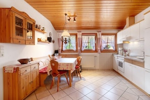 Une cuisine avec des meubles de rangement en bois, une table à manger avec des chaises, un luminaire suspendu et une fenêtre avec des rideaux.