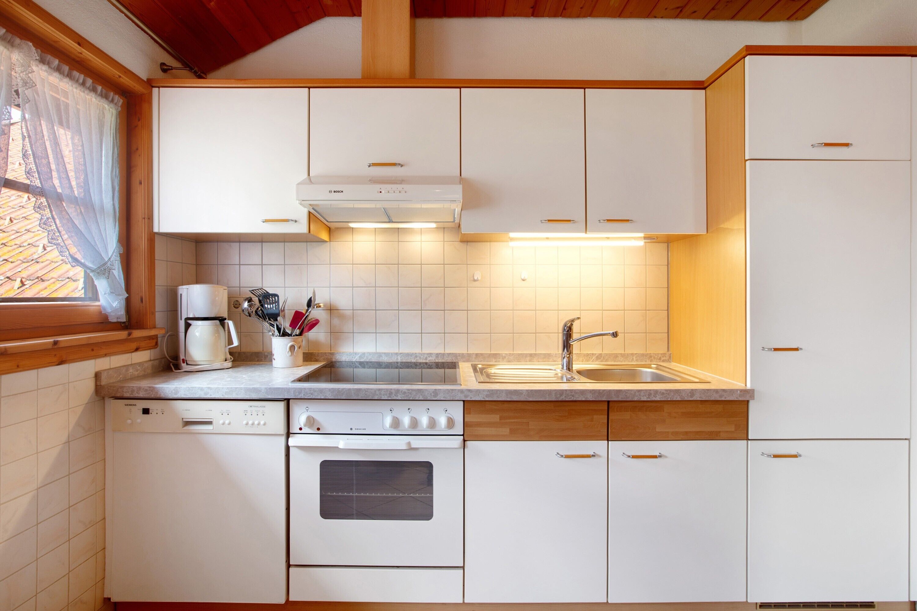 Een keuken met witte kastjes, een vaatwasser, een oven, een spoelbak en een raam met gordijn.
