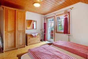 Une pièce avec un plancher en bois, une armoire en bois, une commode, un miroir, deux lits avec une literie à carreaux et une fenêtre avec des rideaux.