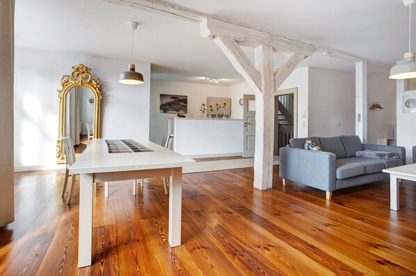 A spacious room with a wooden floor, a white table, a grey sofa, and a large ornate mirror.