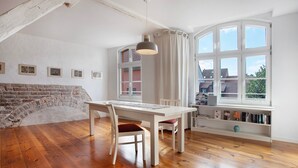 Ein Essbereich mit einem weißen Tisch und Stühlen, einer Hängeleuchte, einer Ziegelwand und einem Fenster mit Blick auf Gebäude.