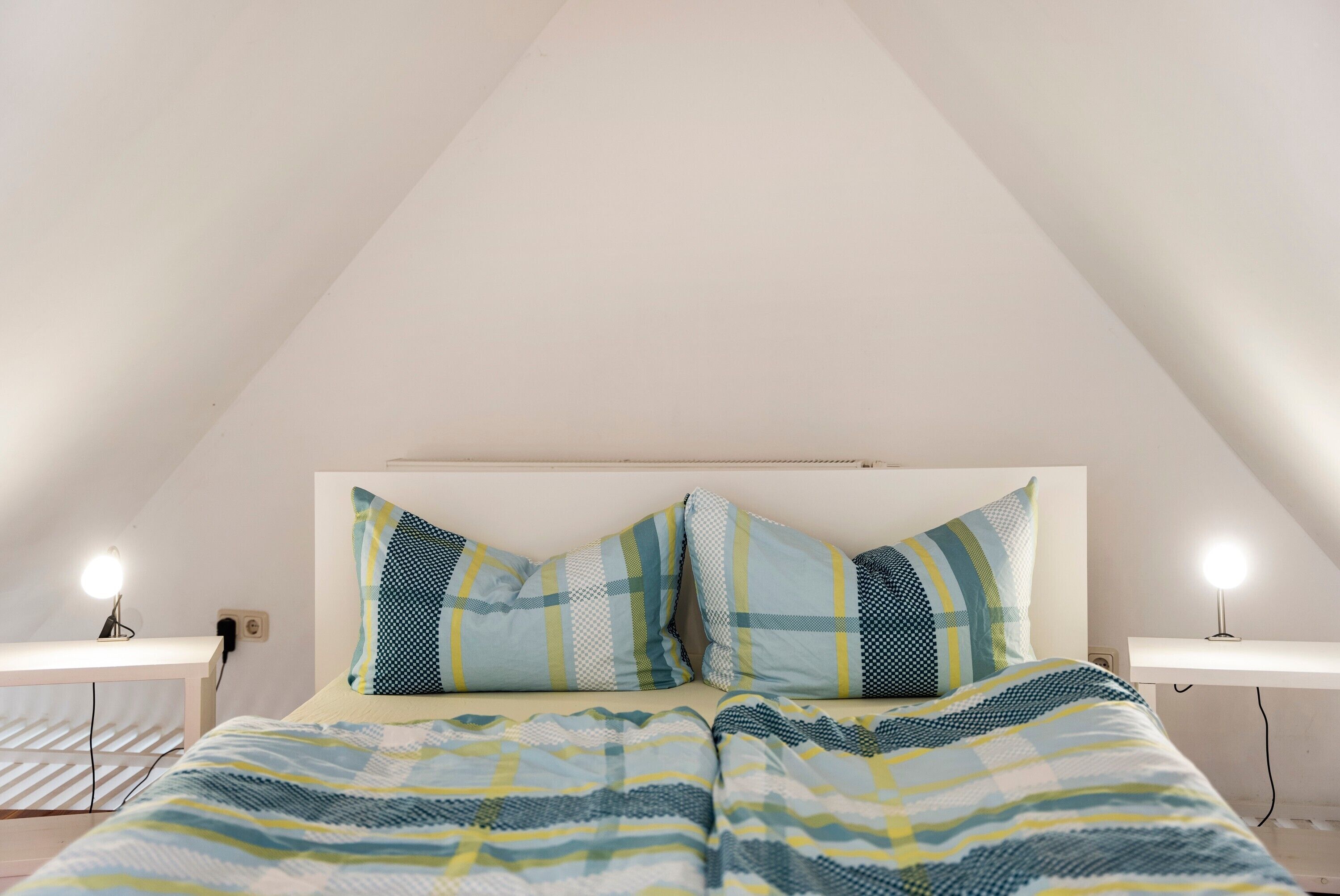 Ein Schlafzimmer mit schrägen Wänden, einem Bett mit gemusterten Betttextilien, zwei Nachttischlampen und einem weißen Kopfteil.