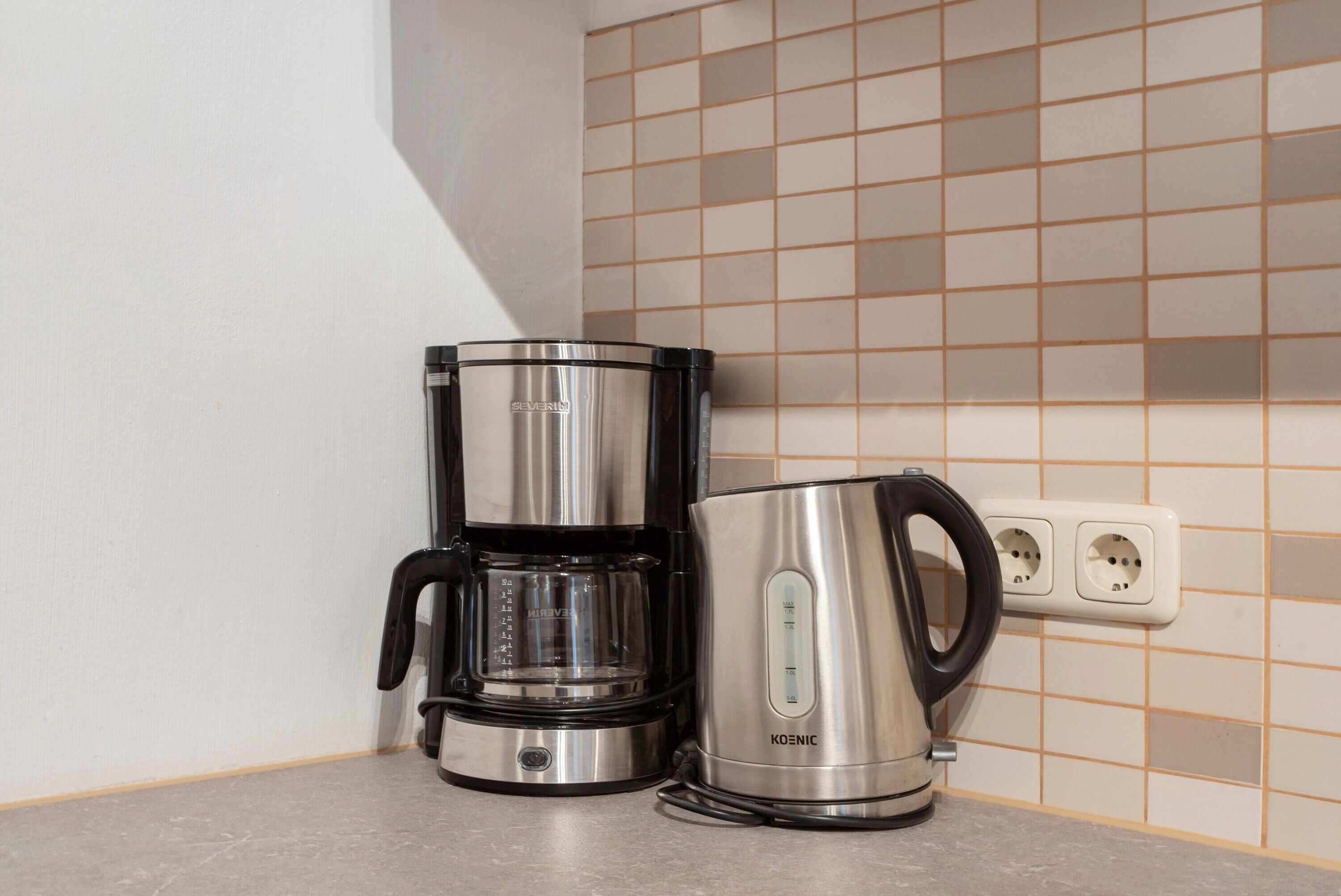 A stainless steel coffee maker and electric kettle on a kitchen counter with a tiled backsplash.