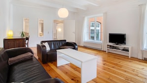 Ein modernes Wohnzimmer mit einem Ledersofa, einem weißen Couchtisch, einem Fernseher auf einem Standfuß und einem großen Fenster mit Vorhängen.