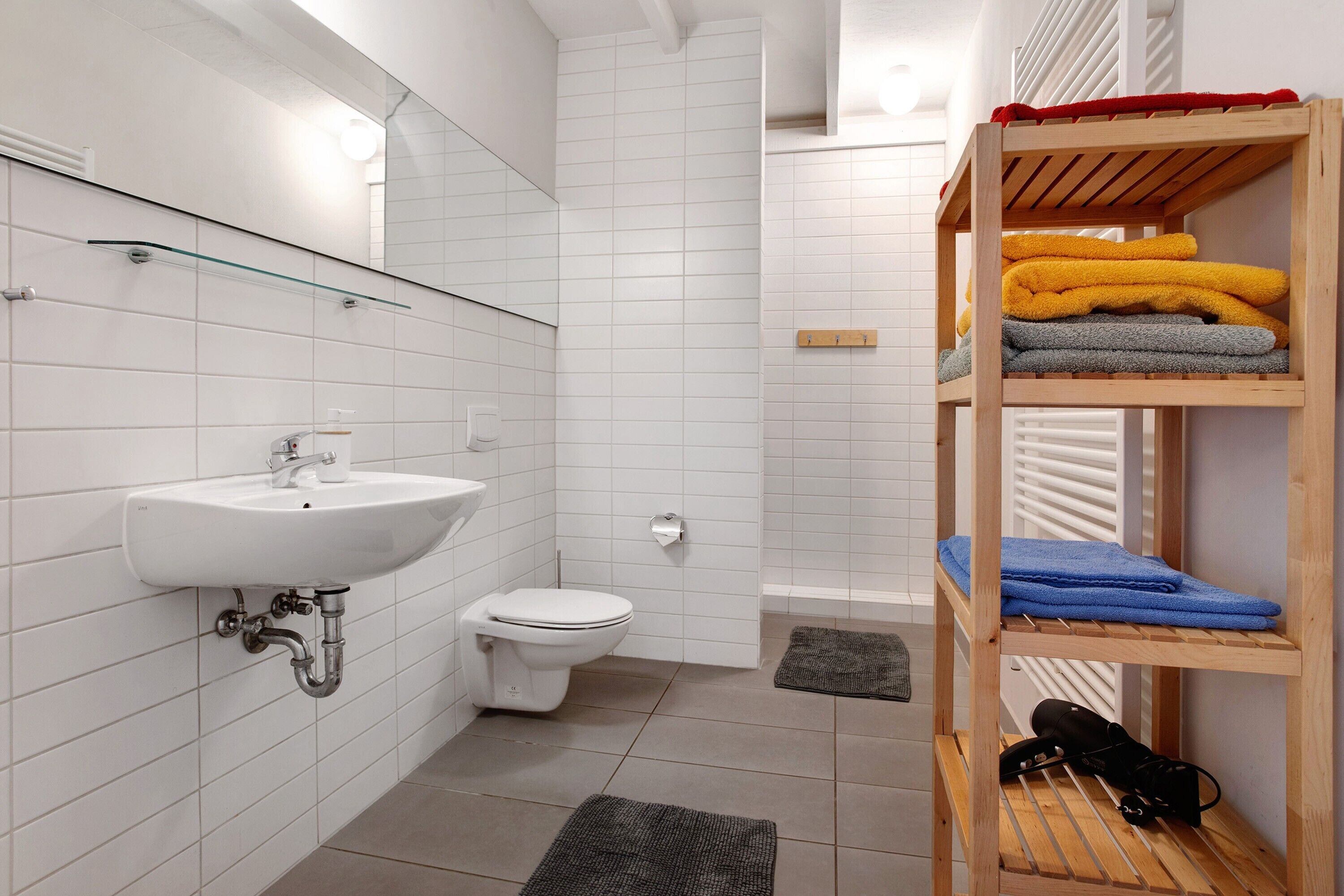 A bathroom with a sink, toilet, and shower. There is a shelf with towels and a bag.