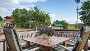 Una terraza de madera con una mesa y sillas, una maceta con flores moradas, y una vista de casas y árboles al fondo.
