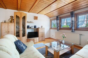 Ein gemütliches Wohnzimmer mit Holzboden, einer Couch, einem Glastisch, einem Fernseher und einem Fenster mit Vorhängen.