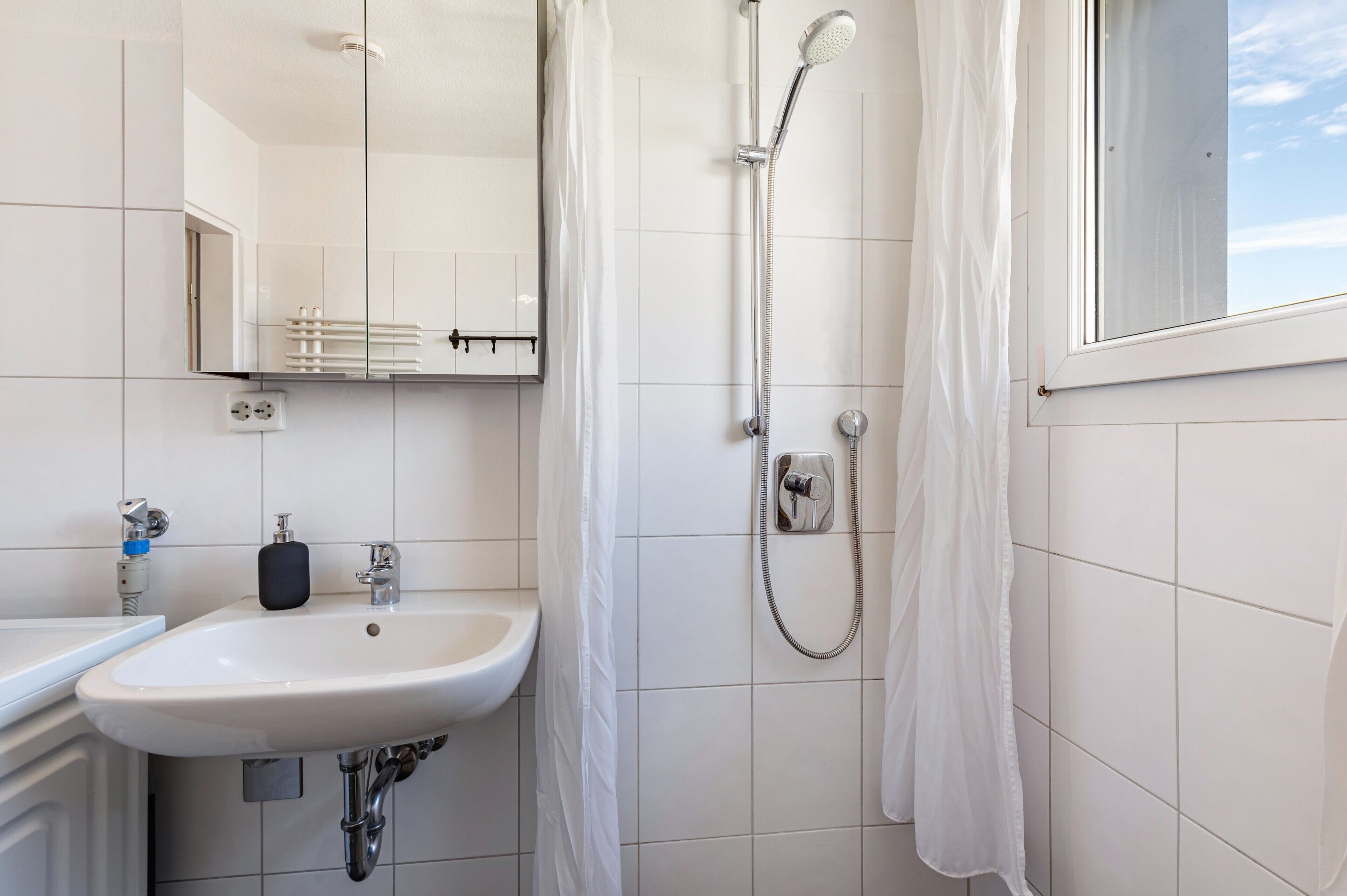 Un baño con lavamanos blanco, un espejo, ducha con cortina y una ventana.