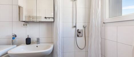 Un baño con lavamanos blanco, un espejo, ducha con cortina y una ventana.