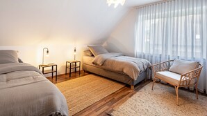 Ein Schlafzimmer mit Bett, Stuhl, Nachttisch, Lampe und einem Fenster mit Vorhängen.