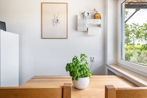 A kitchen with a wooden table, a potted basil plant, a framed picture, and a window with a view of greenery.