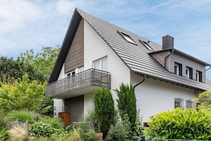 A modern two-story house with a sloped roof, a balcony, and a garden with various plants.