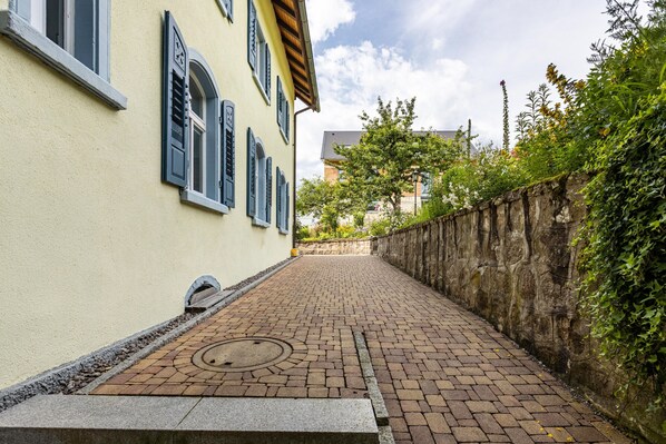 A paved pathway leading to a building with a stone wall on one side and a tree-lined street on the other.