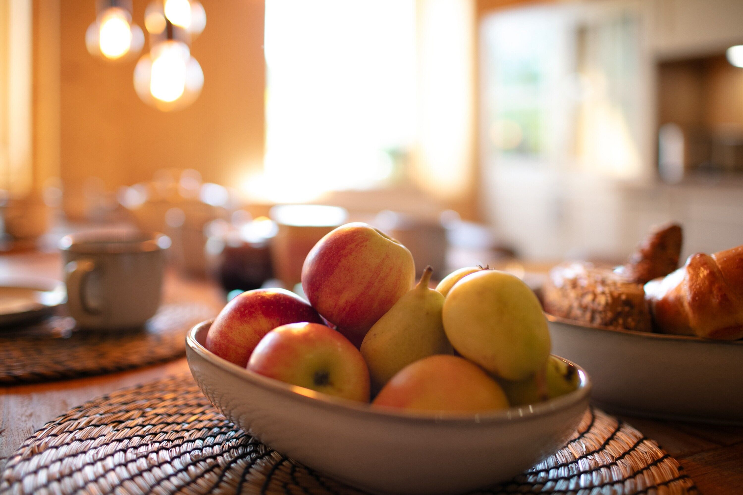A bowl of fruit on a table with a woven placemat, a cup, and a croissant in the background.