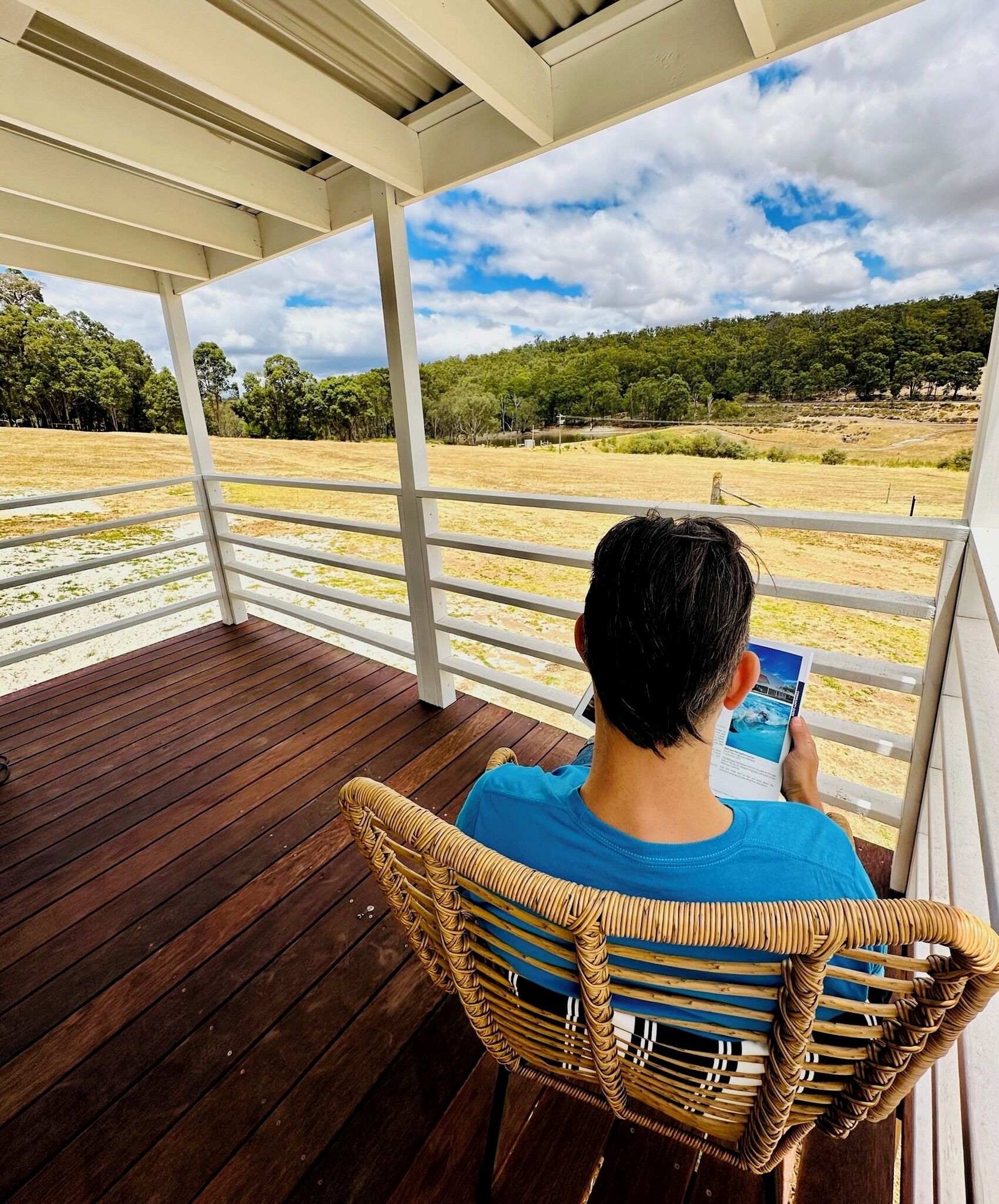 A person sitting on a wooden deck chair under a white pergola, reading a book.
